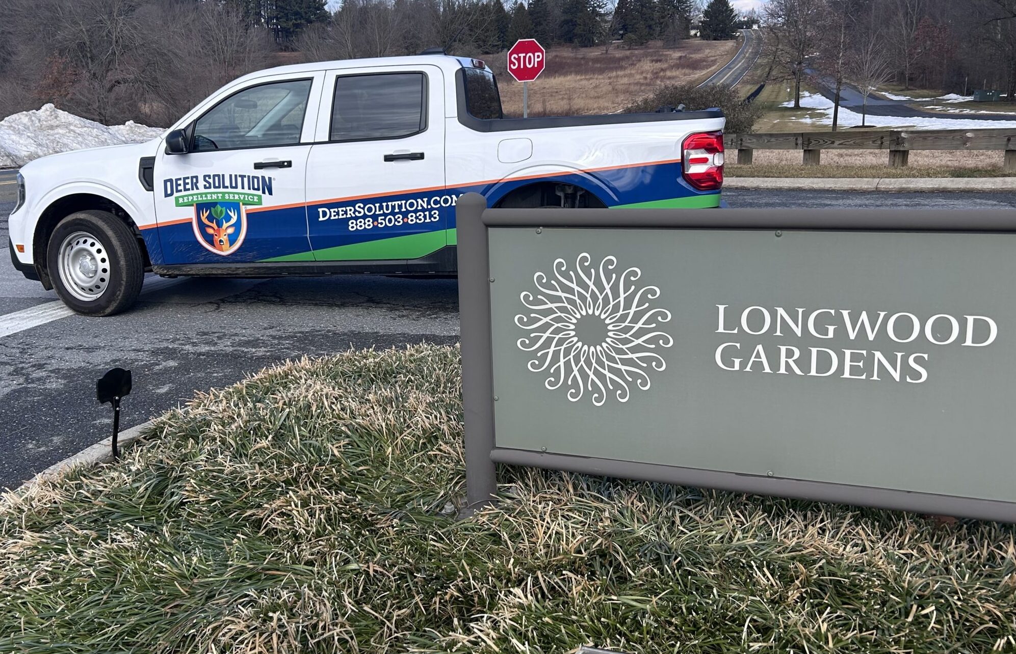 White pickup truck with Deer Solution branding parked near a Longwood Gardens sign in a winter landscape.