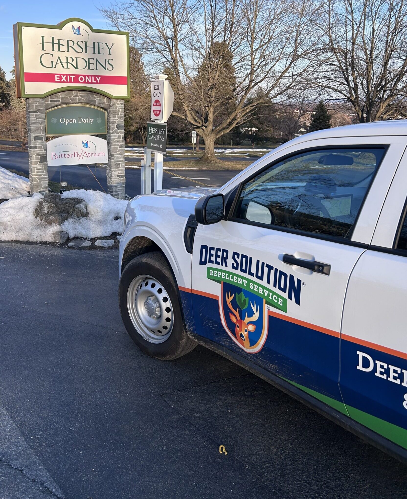 White Deer Solution pest control truck parked beside a Hershey Gardens exit sign, with snow piles and bare trees in a winter lot.