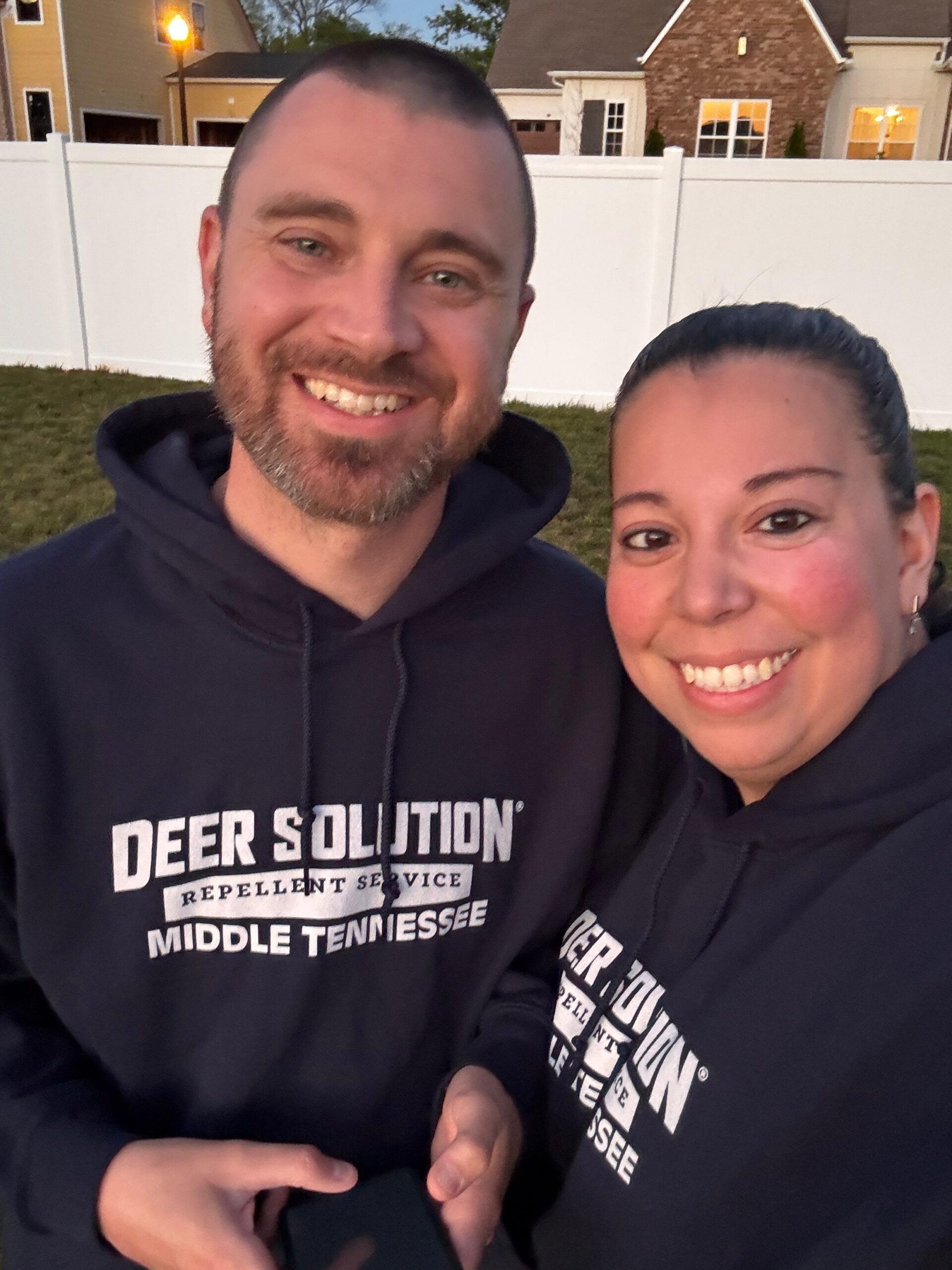 Two people smiling for a selfie outdoors in front of a white fence; both wear black hoodies with a Deer Solution logo and text mentioning Tennessee.