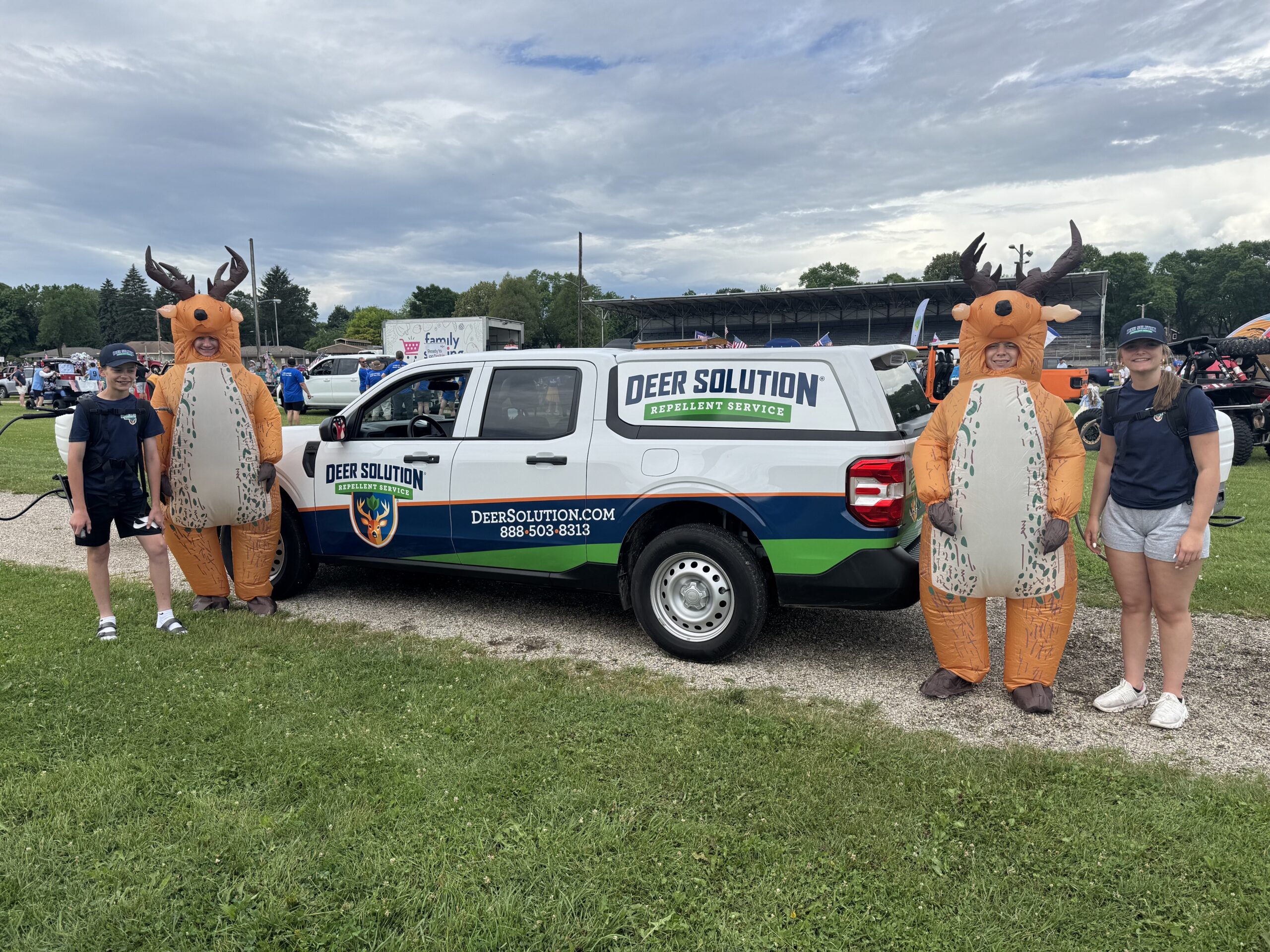 Two people in inflatable deer costumes stand beside a Deer Solution repellent service van at an outdoor event ground, with others in the background.