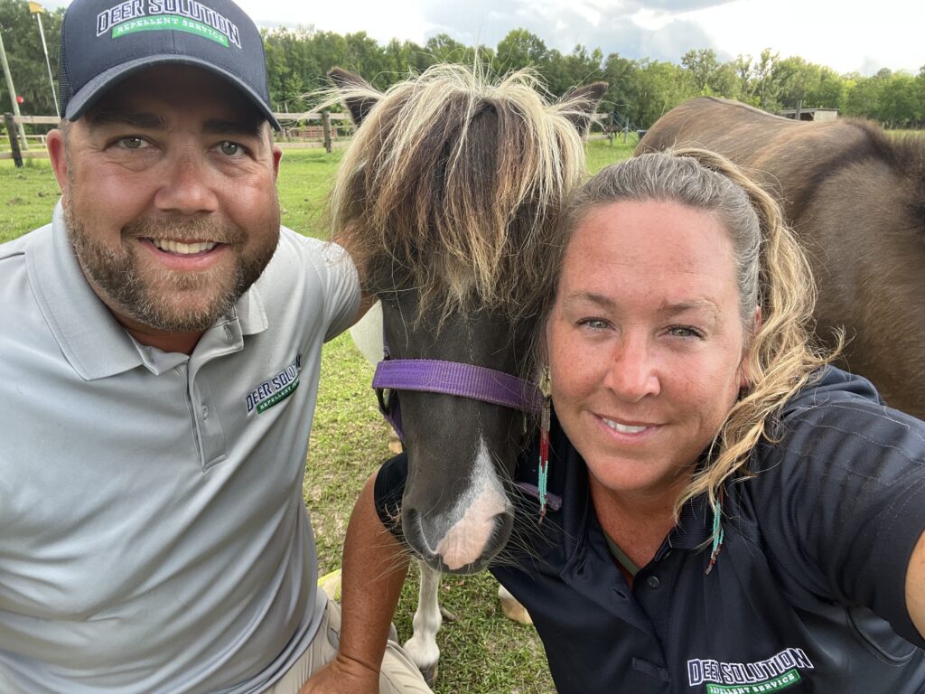 Two people taking a close selfie with a brown pony between them in a grassy outdoor area; man wearing a cap, woman in dark shirt.