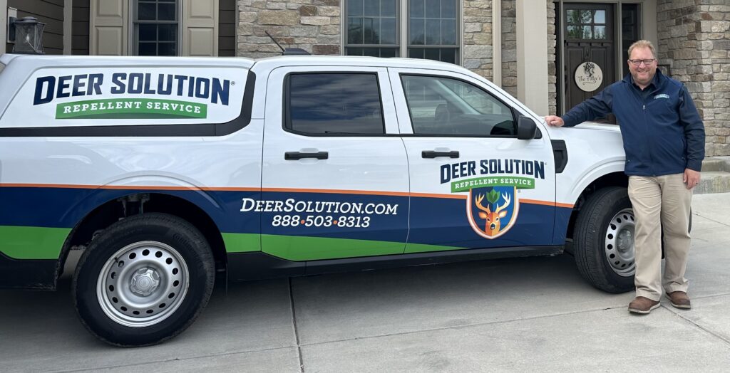 Man in a blue jacket stands beside a Deer Solution Repellent Service van parked in front of a house exterior.