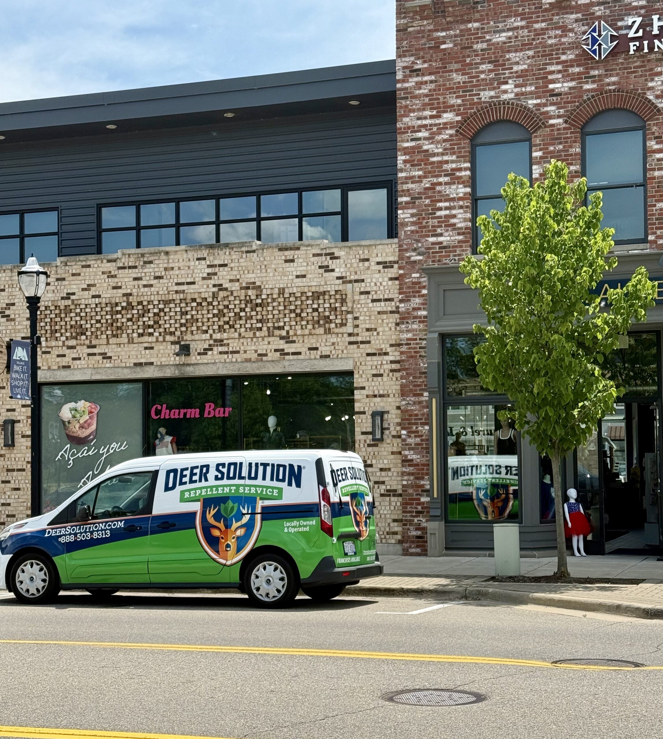 Pest-control van labeled 'DEER SOLUTION' parked in front of a brick storefront with a 'Charm Bar' sign and a leafy green tree nearby.