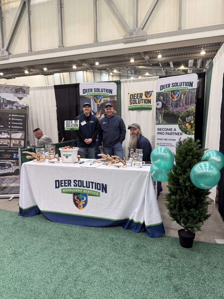 Trade show booth for Deer Solution Repellent Service with three men behind a table, banners, and teal balloons.