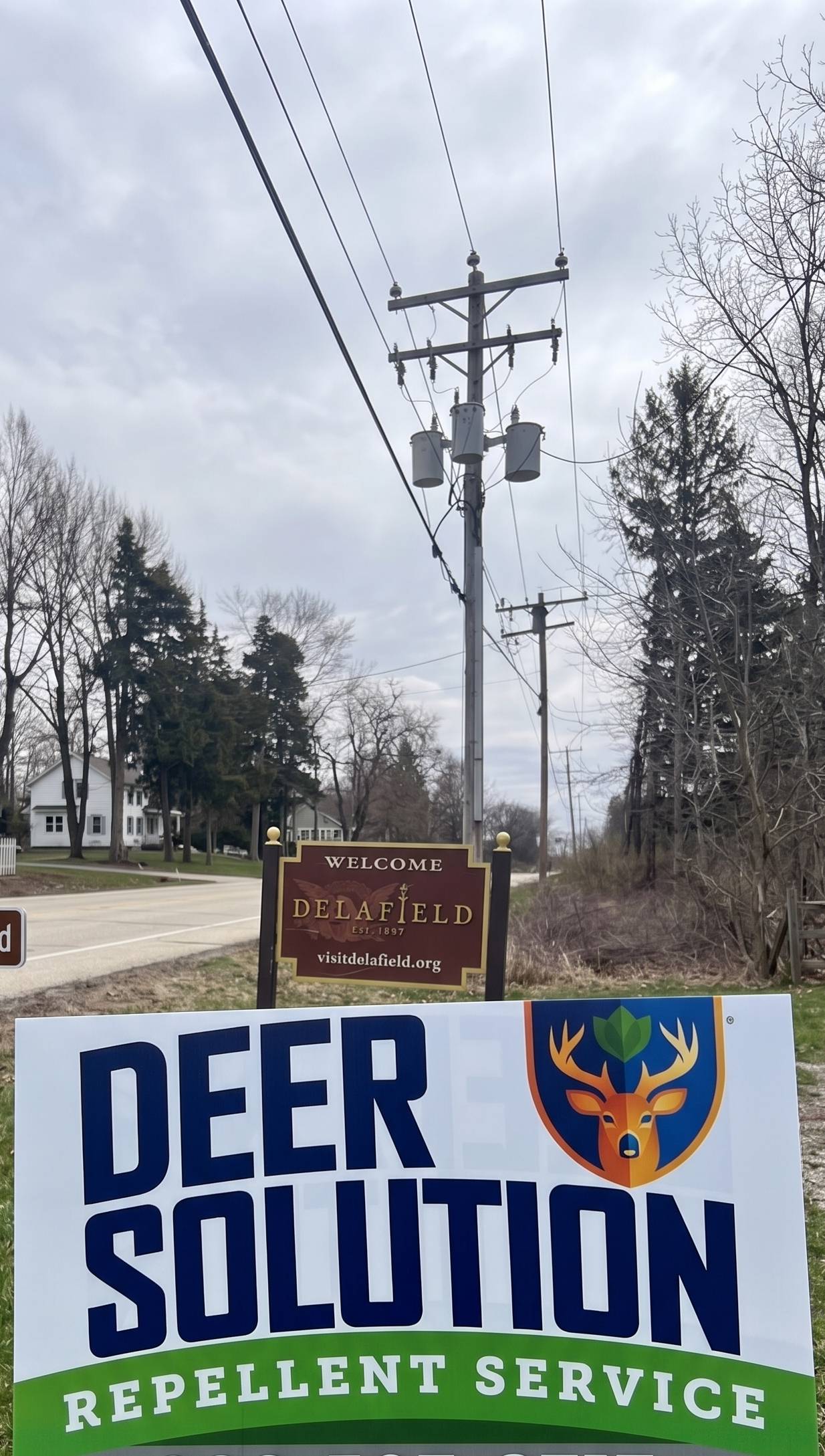 Public roadside scene with a utility pole and two welcome signs: Delafield sign and a deer repellent service sign nearby, in a suburban area.