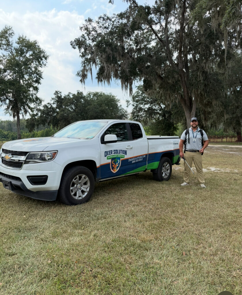 Pest control technician in uniform standing beside a branded white pickup truck in a grassy park area with trees overhead.