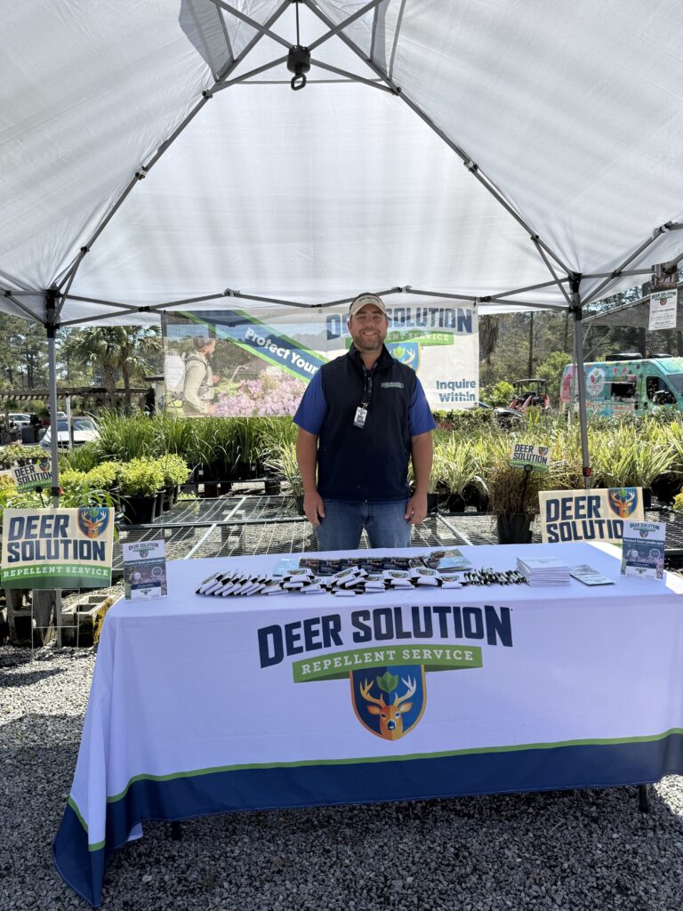 Man in a blue vest stands under a canopy at a Deer Solution promo booth with a table of flyers and pens showing deer logo signs behind him.