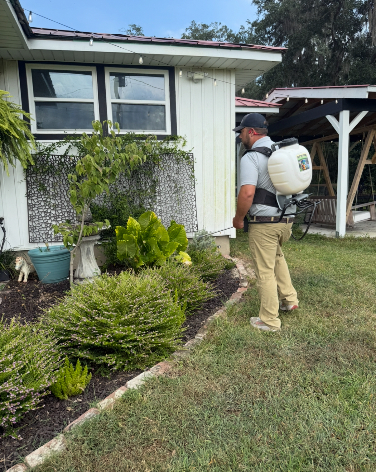 Man in cap with a backpack sprayer spraying plants along a garden bed beside a house.