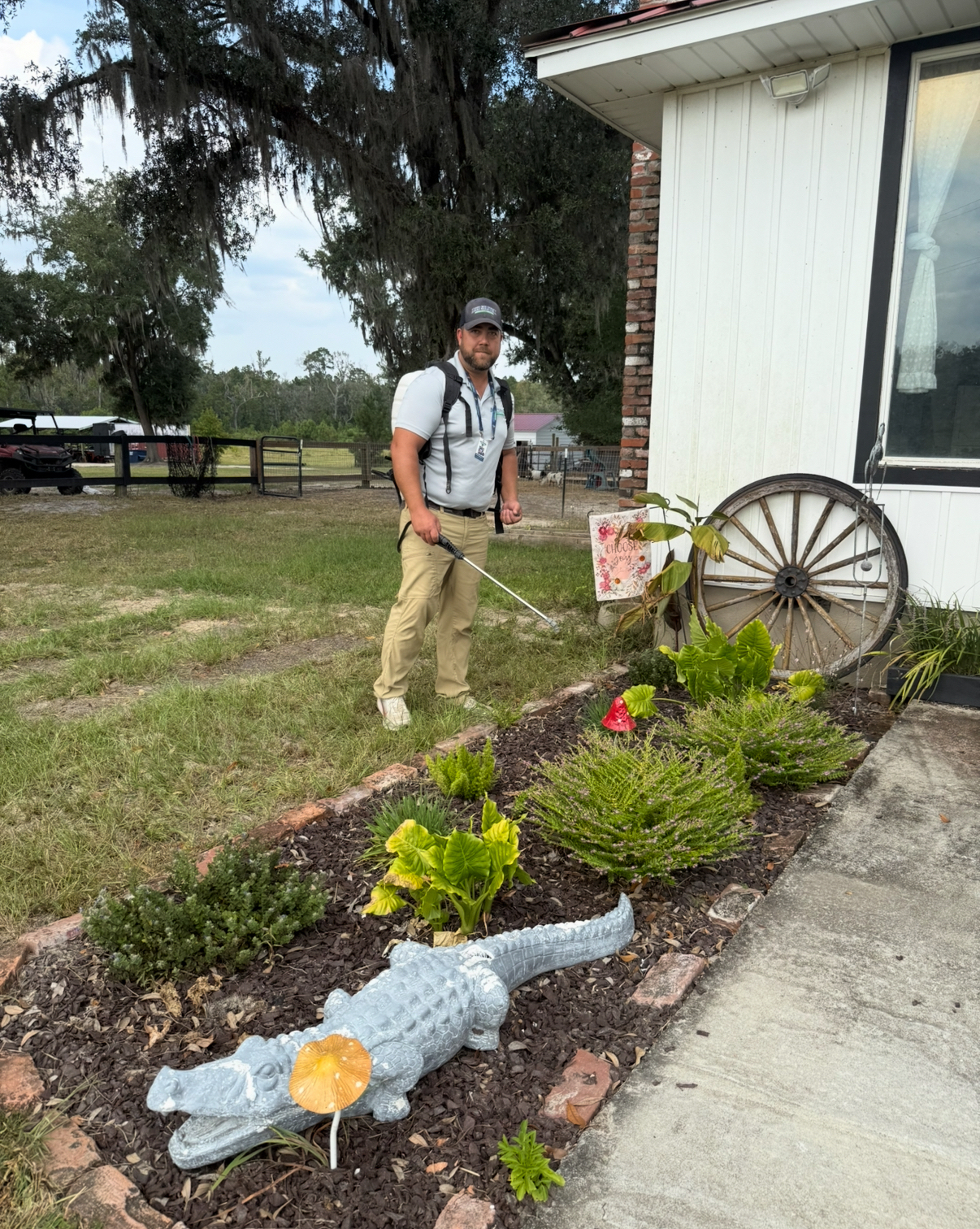 Man with a backpack sprayer standing over a garden bed outside a white house with a wagon wheel decor and various plants.