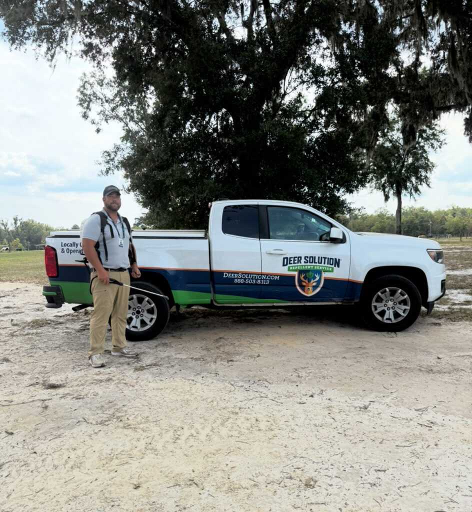 Man in light shirt and cap stands beside a white Deer Solution service truck on a dirt lot under a tree edge.