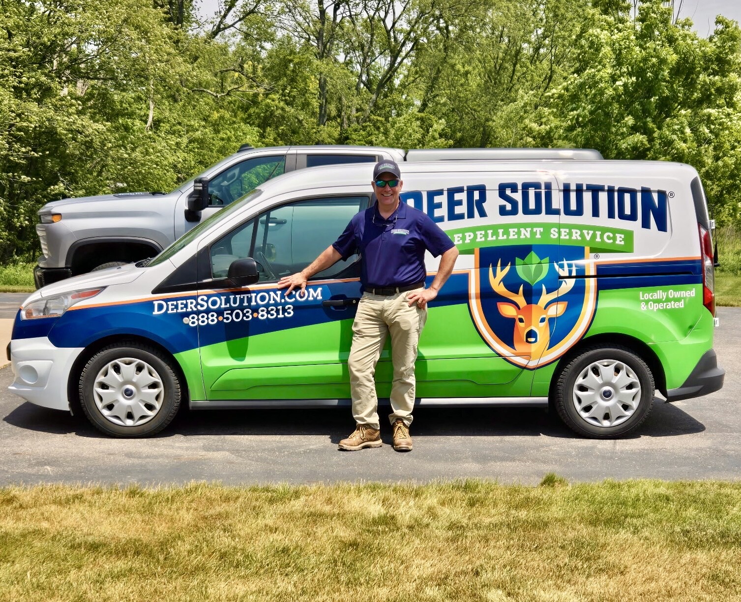 A man in a navy polo and cap stands beside a colorful Deer Solution pest control van with a deer logo in a grassy area.