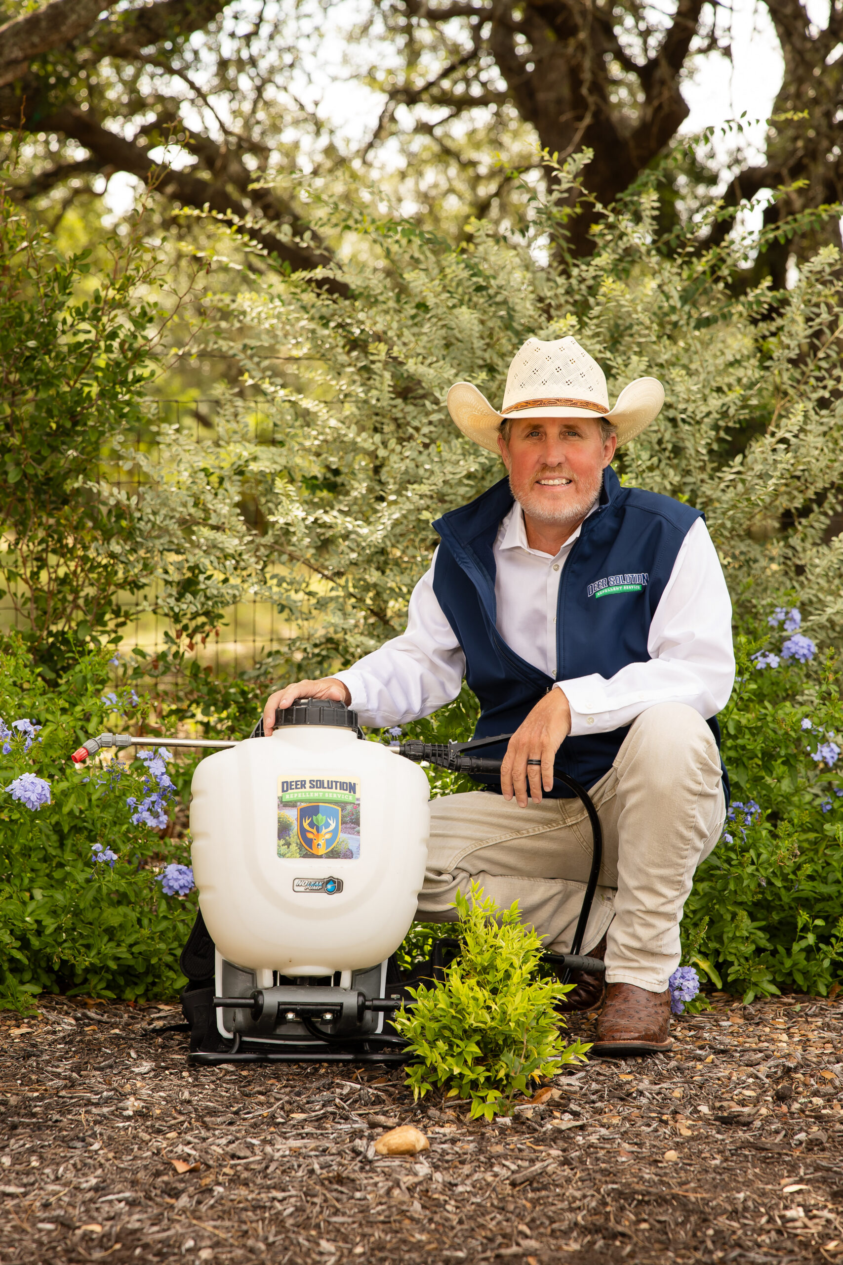 Man in a cowboy hat kneeling in a garden beside a white sprayer, ready to apply liquid to plants.